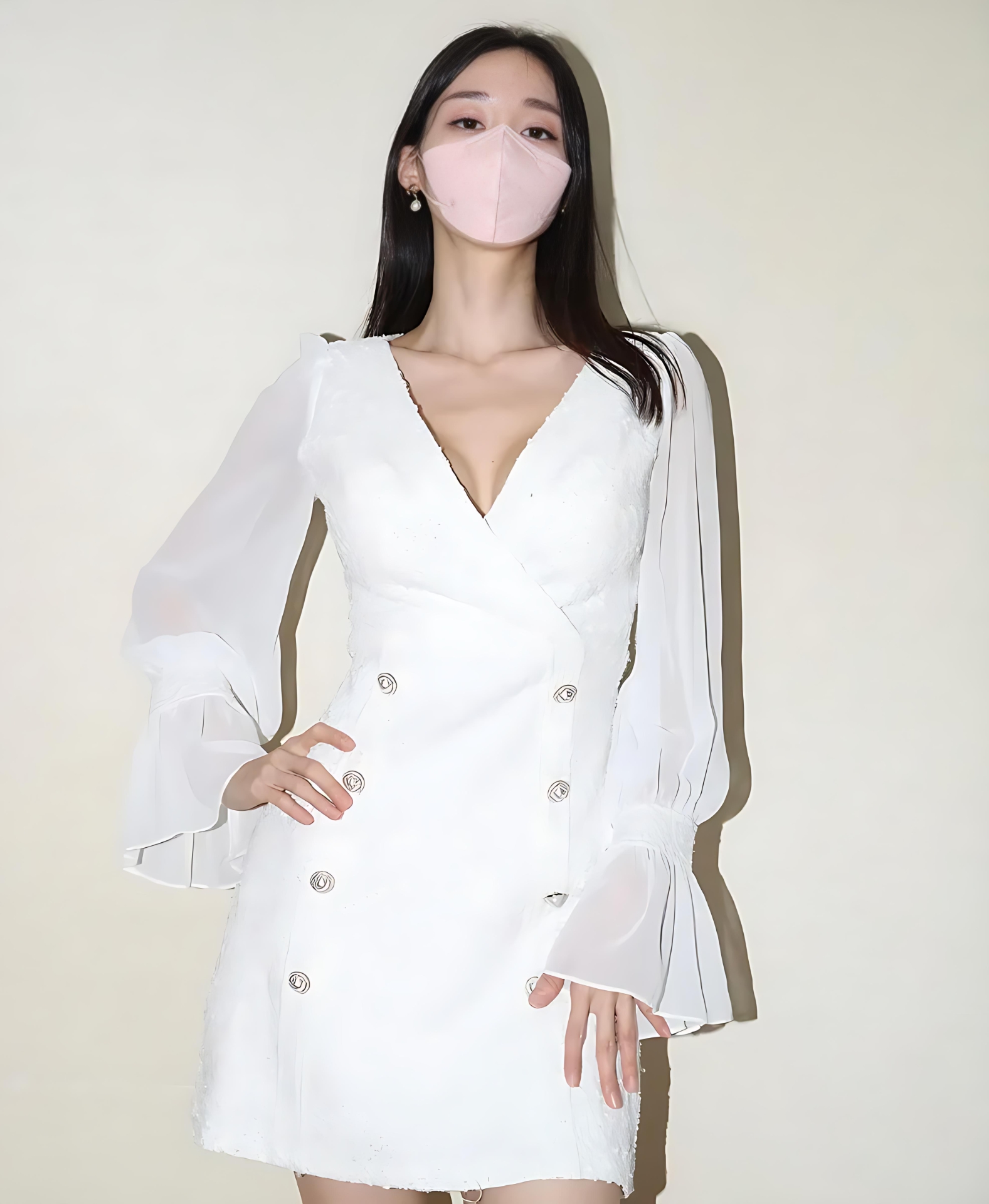 公开 长黑发人物佩戴粉色口罩，穿白色长袖连衣裙站在浅色墙前