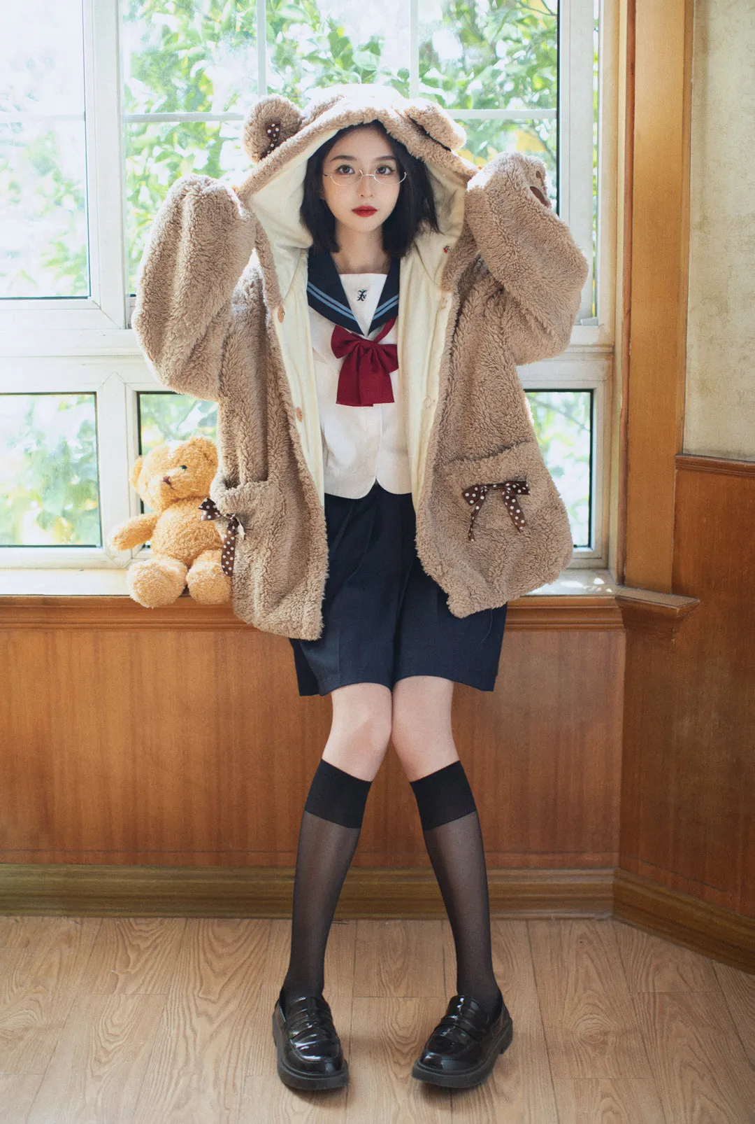 女生穿棕色毛绒连帽外套和水手服站在窗前,双手抬起扶住帽子
