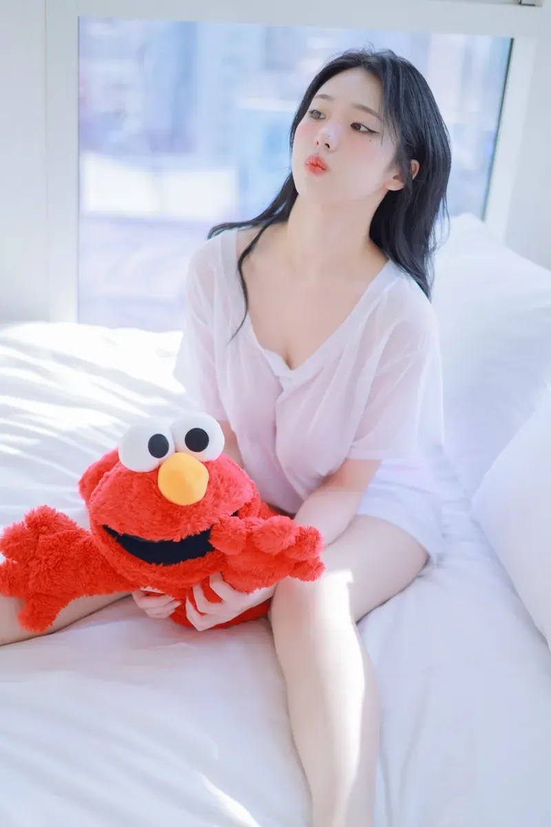 清月已经不困了身着白色透气短袖上衣在纯白床单上抱红色艾摩玩偶端坐近景特写