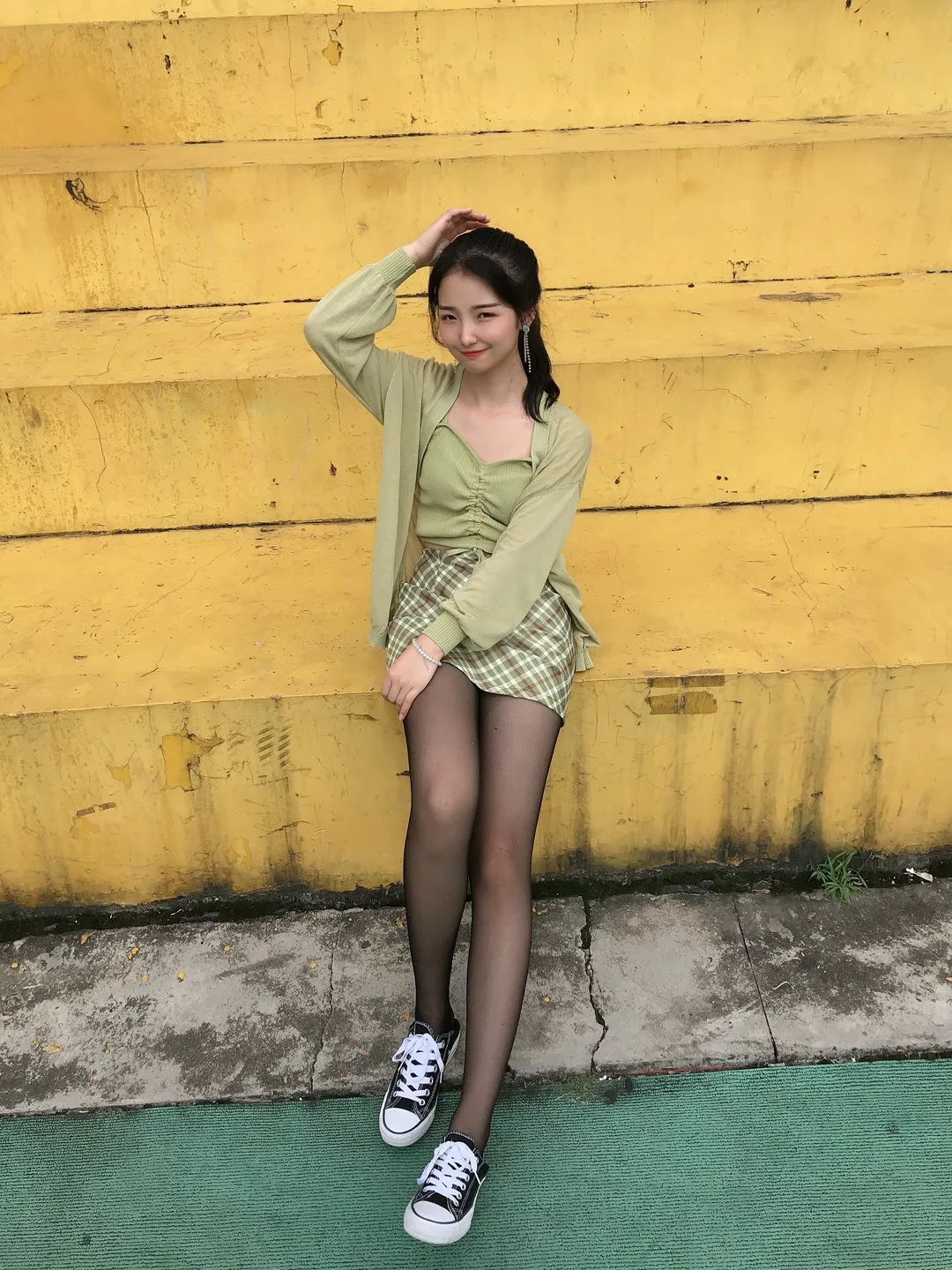 黄色斑驳墙壁浅草绿针织开衫嫩绿吊带格纹裙黑丝配经典帆布鞋立姿